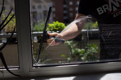 Nahaufnahme einer Person, die ein Fenster mit einem Abzieher reinigt, mit Wassertropfen und Seife auf der Scheibe, sonniger Tag draußen