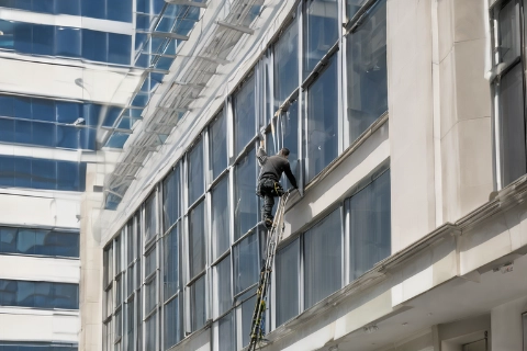 Ein Gebäudereiniger in Sicherheitsausrüstung reinigt die Fenster eines Hochhauses, gesichert durch ein Seilsystem